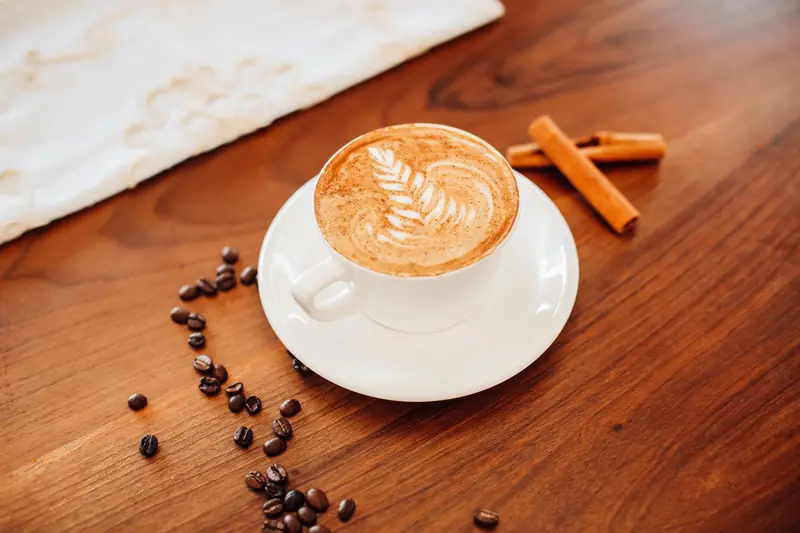 A cup of cappuccino with intricate latte art on a wooden table, accompanied by coffee beans and cinnamon sticks