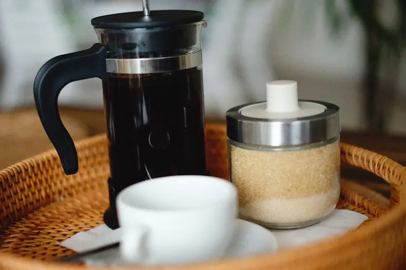 Close-up of a French press, sugar jar, and coffee cup on a wicker tray