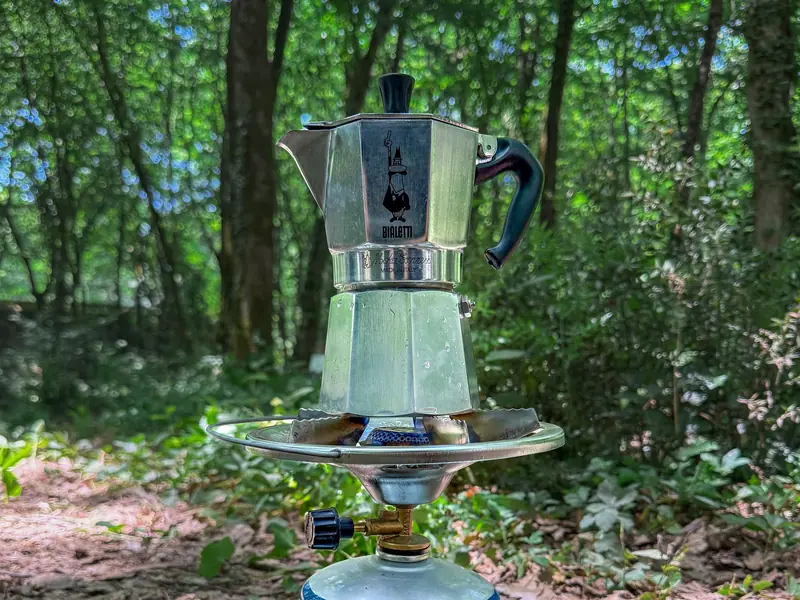 Moka pot brewing coffee on a portable stove amidst forest greenery, capturing a serene outdoor experience